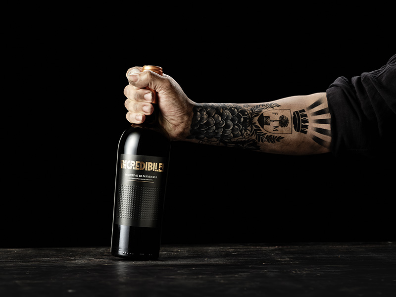 Primitivo di Manduria aus Apulien iNCREDIBILE! Mit Trauben tätowierter Arm hält eine Flasche iNCREDIBILE! - Primitivo di Manduria aus Apulien