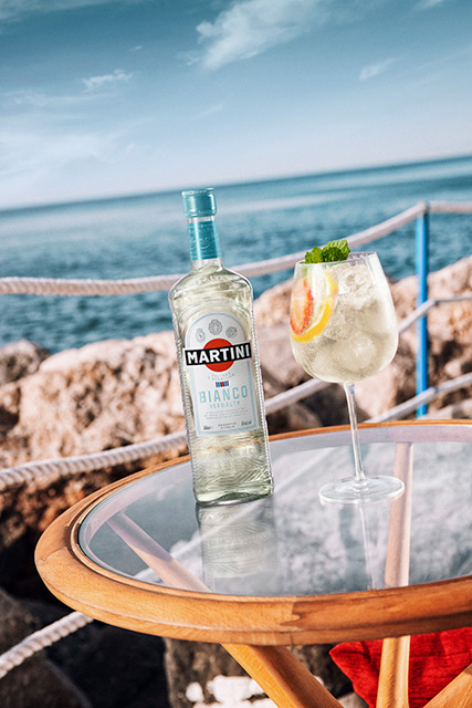 MARTINI Bianco Spritz bei GH (1) Ein MARTINI Bianco Spritz im Glas mit einer Flasche MARTINI Bianco, serviert direkt am Meer