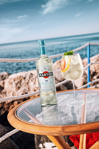 Ein MARTINI Bianco Spritz im Glas mit einer Flasche MARTINI Bianco, serviert direkt am Meer
