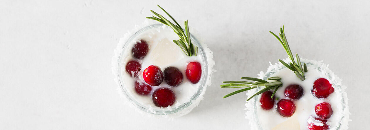 Ein köstlich cremiger Genuss: unser Cheesecake-Likör, hübsch dekoriert mit Cranberrys, Rosmarin und einem Rand aus Kokosraspeln.